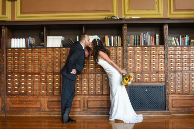 The bride and groom steal a kiss in front of the catalog shelves.